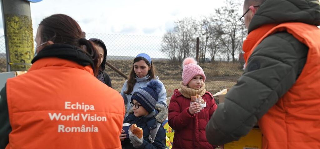 Three children wearing winter coats and hats and holding baked goods and treats face two World Vision staff members in heavy orange vests, a woman on the left and a man on the right.