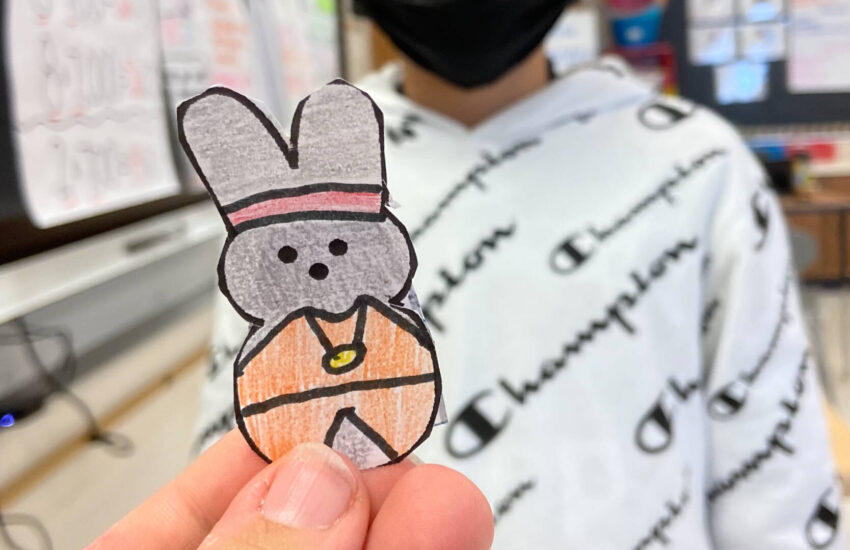 A woman’s hand holds a cutout and hand-colored drawing of a Peep in an orange cape. It’s how one of her students sees her.